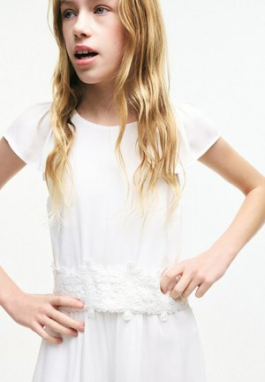 Blondynka z falującymi włosami, ubrana w białą sukienkę z kwiatowym koronkowym paskiem, z rękami na biodrach, spoglądająca lekko na bok.