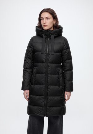 Femme portant un long manteau matelassé noir avec un col montant et une fermeture éclair sur le devant, debout devant un fond blanc uni.