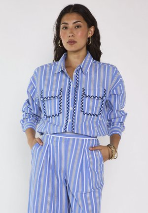 Donna che indossa una camicia a righe verticali blu e bianca con bordi neri e pantaloni a righe coordinati a vita alta, mani in tasca, orecchini a cerchio e braccialetti.