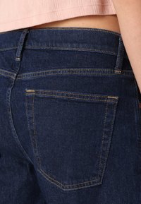 Jeans de mezclilla azul oscuro con una textura suave, que presentan un bolsillo trasero con costuras amarillas y un diseño de cintura media.
