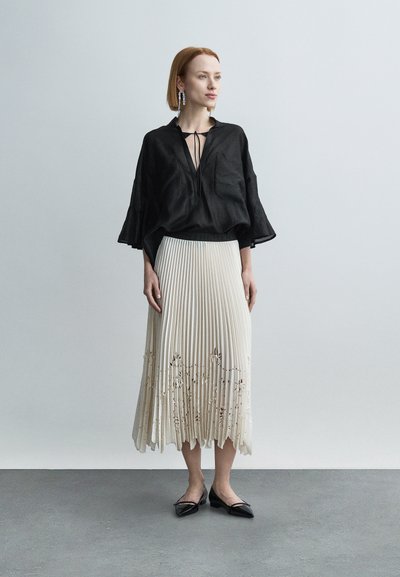 Μαύρο λινό τοπ με φαρδιά μανίκια, σε συνδυασμό με κρεμ πλισέ μίντι φούστα με κεντήματα με λουλούδια και μαύρες μυτερές μπαλαρίνες.