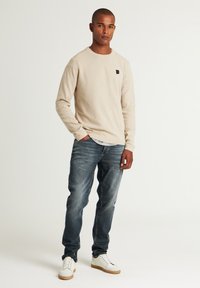 Beige Hemd mit langen Ärmeln und strukturiertem Finish, kombiniert mit abgedunkelten blauen Jeans und weißen Sneakers mit braunen Akzenten. Schwarzes Logo-Detail auf dem Hemd.