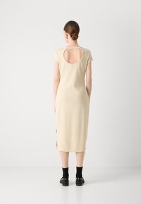 Beige jersey dress with short sleeves, open back, knee-length, side slits, worn with black socks and slip-on shoes. Smooth texture.