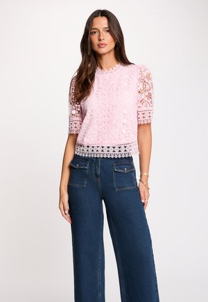 Frau trägt ein rosa Spitzen-Top mit kurzen Ärmeln und komplexen Mustern sowie dunkelblaue Jeans mit weitem Bein und aufgesetzten Fronttaschen.