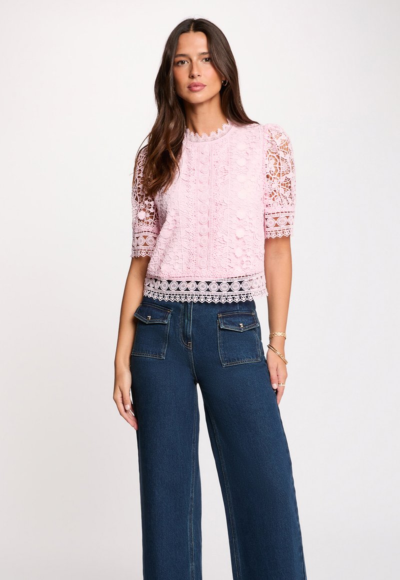 Mujer con una blusa rosa de encaje de manga corta con patrones intrincados y vaqueros anchos azul oscuro con bolsillos delanteros con solapa.