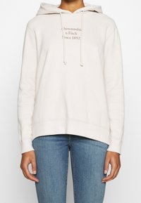 Hellbeiger Hoodie aus weichem Stoff mit einer vorderen Kängurutasche, verstellbaren Zugbändern und bedrucktem Logo-Text. Lockerer Schnitt.