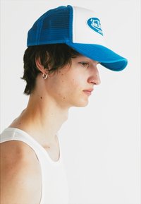 Gorra trucker azul y blanca con lados de malla; presenta un visera curva y una etiqueta redonda en la parte frontal que muestra un logo.
