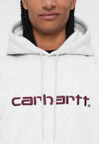 Felpa grigia con finitura testurizzata, caratterizzata da un grande logo "carhartt" ricamato in bordeaux e con coulisse regolabili.