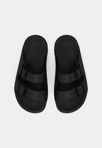 Une paire de sandales noires à glisser avec deux sangles réglables à boucles et des semelles texturées sur un fond blanc.