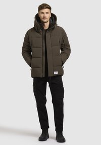 Tamnozeleni pufast kaput s kapuljačom, s kvadratnim panelima i prednjim patentnim zatvaračem, nosi se preko crne majice i uparen je s crnim cargo hlačama.