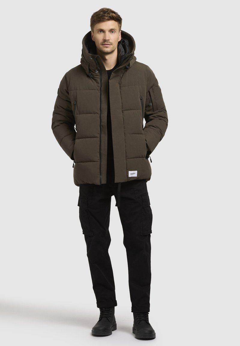Dunkelgrüne gefütterte Jacke mit Kapuze, ausgestattet mit gesteppten Panele und einem Frontreißverschluss, getragen über einem schwarzen Shirt und kombiniert mit schwarzen Cargo-Hosen.