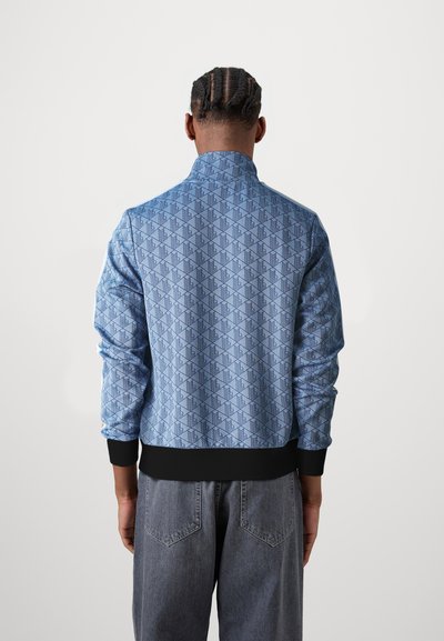 Veste aviateur bleue avec un motif géométrique, col montant, poignets et ourlet côtelés noirs. Fabriquée en tissu lisse avec une texture subtile.