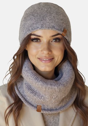 Žena s světlou šedou pletenou čepicí a odpovídajícím silným žebrovaným pleteným návlekem na krk, usmívající se s dlouhými vlnitými hnědými vlasy.