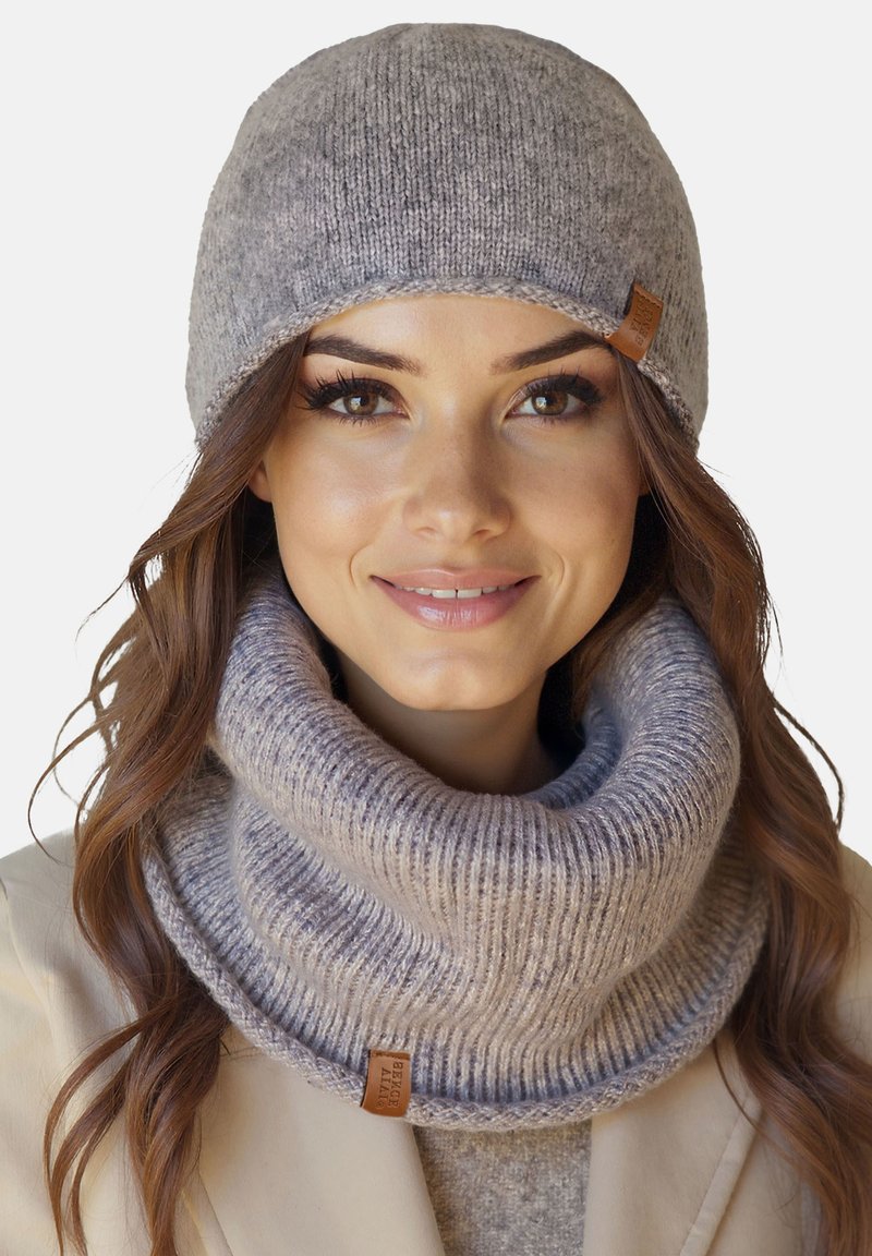 Kvinde iført en lysegrå strikket hat og matchende tyk ribstrikket halsedisse, smilende med langt, bølget brunt hår.