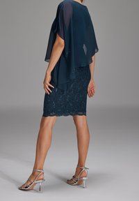 Žena v námořnicky modré krajkové sukni s flitry a průsvitným námořnicky modrým topem s pláštěm, v kombinaci se stříbrnými sandály na vysokém podpatku, stojící.