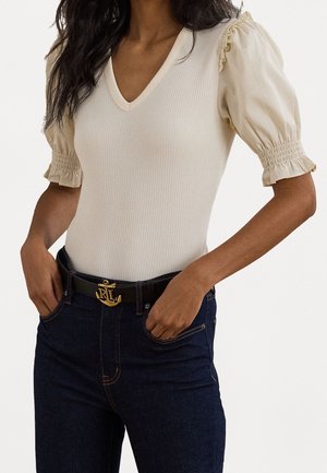 Femme portant un haut crème à col en V avec des manches bouffantes et des poignets smockés, associé à un jean foncé et une ceinture noire avec une boucle d'ancre dorée.