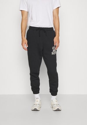 Black sweatpants with elastic cuffs and waistband, featuring a white logo print on the left side. Paired with a white T-shirt and sneakers.