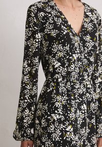 Schwarzes Kleid mit Blumenmuster, das weiße und gelbe Akzente aufweist, einen V-Ausschnitt, lange Ärmel und eine betonte Taille mit einem fließenden Schnitt hat.