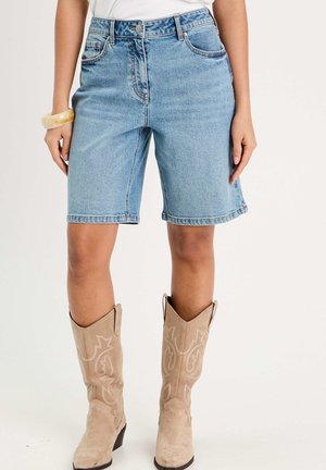 Kvinde iført lyseblå jeansshorts til knæet, beige broderede ruskinds cowboystøvler, hvid top og en beige armbånd.