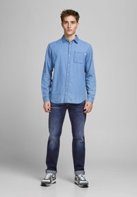 Giovane in piedi, indossa una camicia di denim blu, jeans blu scuro con i polsini arrotolati e sneakers verdi e bianche, su uno sfondo grigio uniforme.