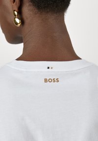 Vue de dos d'une personne portant des boucles d'oreilles en or et une chemise blanche avec "BOSS" imprimé en doré près de l'encolure.