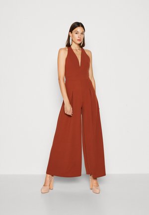 Combinaison couleur rouille avec un décolleté en V profond, des jambes larges et des détails plissés, fabriquée à partir d'un tissu lisse. Complétée par des escarpins à plateforme crème.