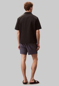 Camisa negra de manga corta, color sólido, con diseño de cuello; combinada con pantalones cortos gris oscuro y sandalias negras, vista desde atrás contra un fondo gris.