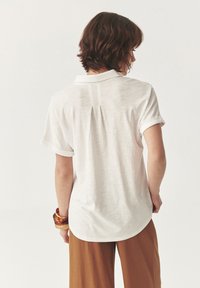 Witte polo met korte mouwen en een ontspannen pasvorm, voorzien van een rugplooi en opgerolde mouwen, gecombineerd met bruine wijde broek.