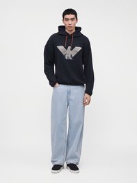 Jeune homme portant un sweat à capuche noir avec un grand logo d'aigle et un jean bleu clair à jambes larges, debout sur un fond blanc uni.