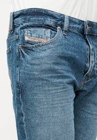 Κοντινό πλάνο μπλε τζιν με εμφανή ετικέτα Diesel Industry στην μικρή μπροστινή τσέπη και χαλκούς κοχλίες στις ενώσεις.