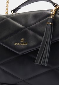 Musta tikattu käsilaukku tekonahkaa, jossa on kullanväriset metalliosat, hapsukoriste ja kohokuvioitu logo. Sisältää ketjuremmin.