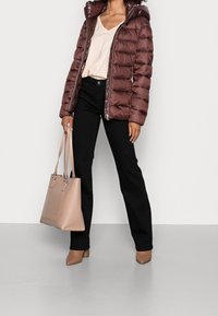 Gepolsterte burgunderrote Jacke mit Kapuze, kombiniert mit einem hellen Oberteil und schwarzen Hosen. Hält eine weiche beige Tasche. Braune Schuhe mit Absatz runden den Look ab.
