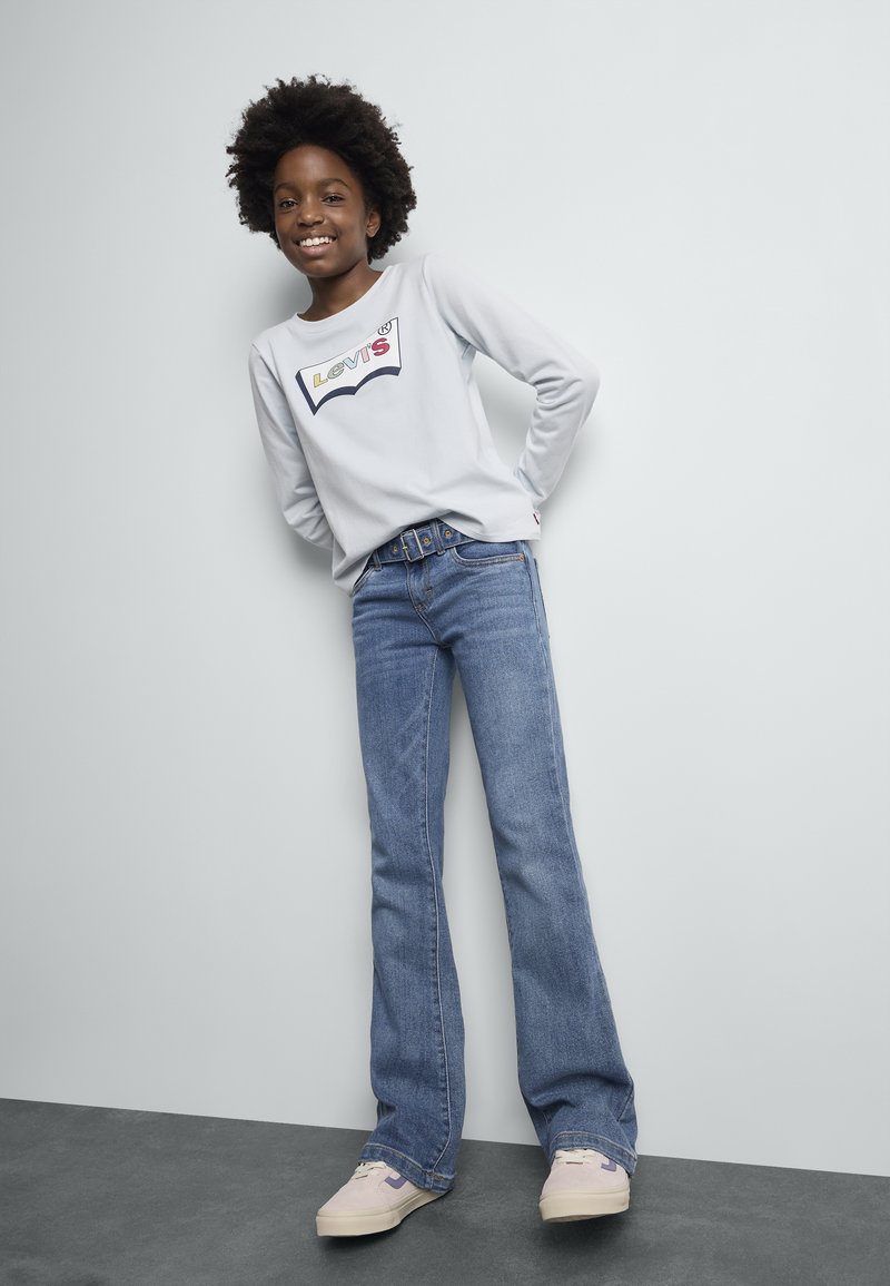 Niño sonriente con peinado afro, vistiendo una camiseta blanca de Levi's de manga larga, jeans azules con cinturón y zapatillas rosa claro, de pie contra una pared blanca.
