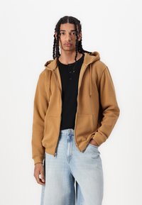 Sweatshirt à capuche zippé en polaire marron, associé à un haut henley noir et un jean large bleu clair. Dispose de poches kangourou.