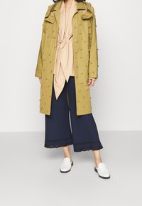 Personne portant un manteau long olive avec des embellissements en 3D, un chemisier beige noué, un pantalon large coupé bleu marine et des mocassins blancs.
