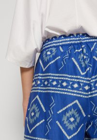 Shorts bleus à motifs avec une taille élastique. Présente des dessins géométriques blancs et des motifs tribaux. Texture de tissu lisse.