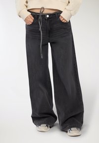 Persona che indossa jeans neri a gamba larga con una cintura annodata e sneakers bianche, con le mani nelle tasche anteriori, su uno sfondo bianco.