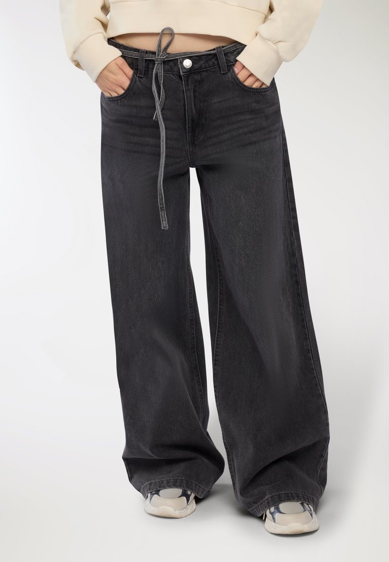 Persona che indossa jeans neri a gamba larga con una cintura annodata e sneakers bianche, con le mani nelle tasche anteriori, su uno sfondo bianco.