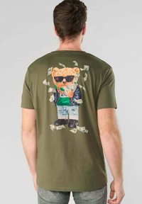Hombre vestido con una camiseta verde oliva que presenta un gráfico de un oso con gafas de sol, chaqueta y pantalones cortos, sosteniendo dinero con billetes flotando alrededor.