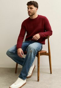 Burgundowa dzianinowa sweter z prążkowanymi mankietami, połączony z przetartymi jasnoniebieskimi dżinsami i białymi sneakersami z brązowymi akcentami, siedzący na drewnianym krześle.