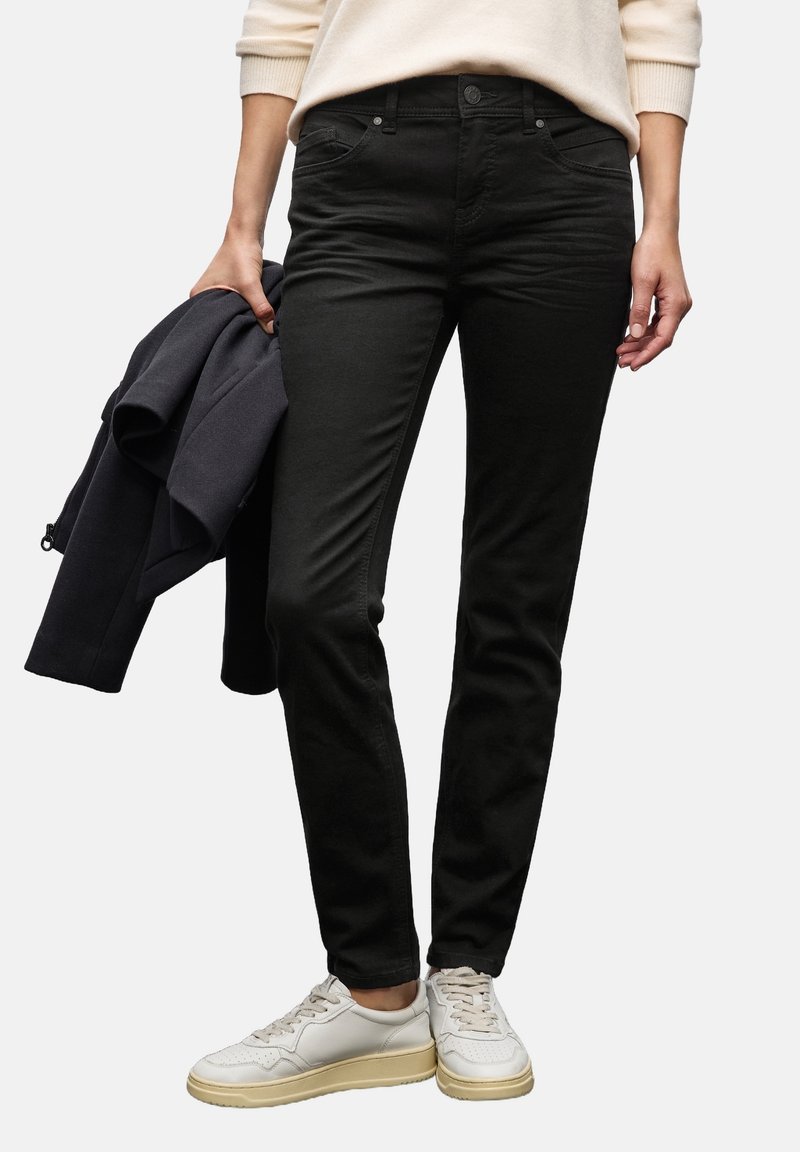 Personne portant un pantalon noir ajusté et des baskets blanches, tenant une veste sombre pliée dans la main gauche.