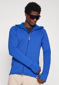 Sudadera azul con cremallera y capucha, hecha de una tela suave y elástica. Cuenta con agujeros para los pulgares y un bolsillo lateral con cremallera, con costuras planas.