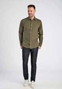 Camisa verde oliva de manga larga con cierre de botones en el frente, un bolsillo en el pecho y un patrón de contraste en el collar interior, combinada con pantalones vaqueros oscuros y zapatillas blancas.