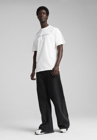 Hvid kortærmet t-shirt med prægede bogstaver, parret med brede sorte jeans og sneakers med hvide detaljer. Glat tekstur, afslappet pasform.