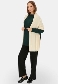 Kremowy szal zarzucony na ciemnozielony sweter, w połączeniu z czarnymi spodniami o szerokiej nogawce i błyszczącymi czarnymi butami. Miękka tekstura i casualowy styl.