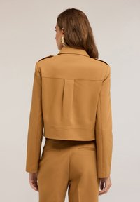 Giacca beige tagliata, con spalle strutturate, taglio dritto e chiusura a bottoni. Tessuto liscio con dettaglio a piega sul retro.