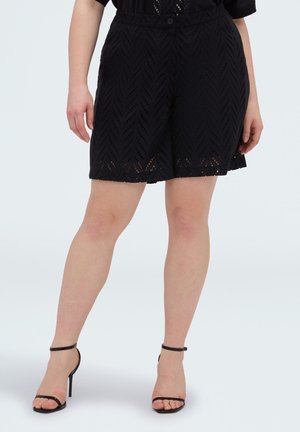 Vrouw draagt zwarte korte broek met patroon en zwarte sandalen met hoge hakken, staand tegen een effen lichte achtergrond.