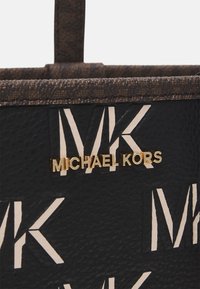 Dettaglio ravvicinato di una borsa nera in stile texturizzato di Michael Kors con motivo beige a logo MK e scritta dorata "Michael Kors" sul davanti.