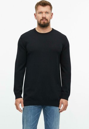 Bearded man wearing a black long-sleeve sweater and blue jeans standing against a plain white background.
