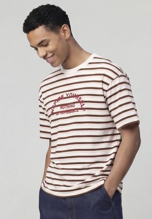Jeune homme souriant, portant un T-shirt rayé marron et blanc avec un texte rouge « INSPIRE YOURSELF, NOTHING IS IMPOSSIBLE », mains dans les poches.